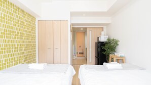 A hotel room with two beds, a wooden wardrobe, a small table with a plant, and a kitchenette in the background. - Shinsaibash East Nana Apartment · Junan (Osaka)