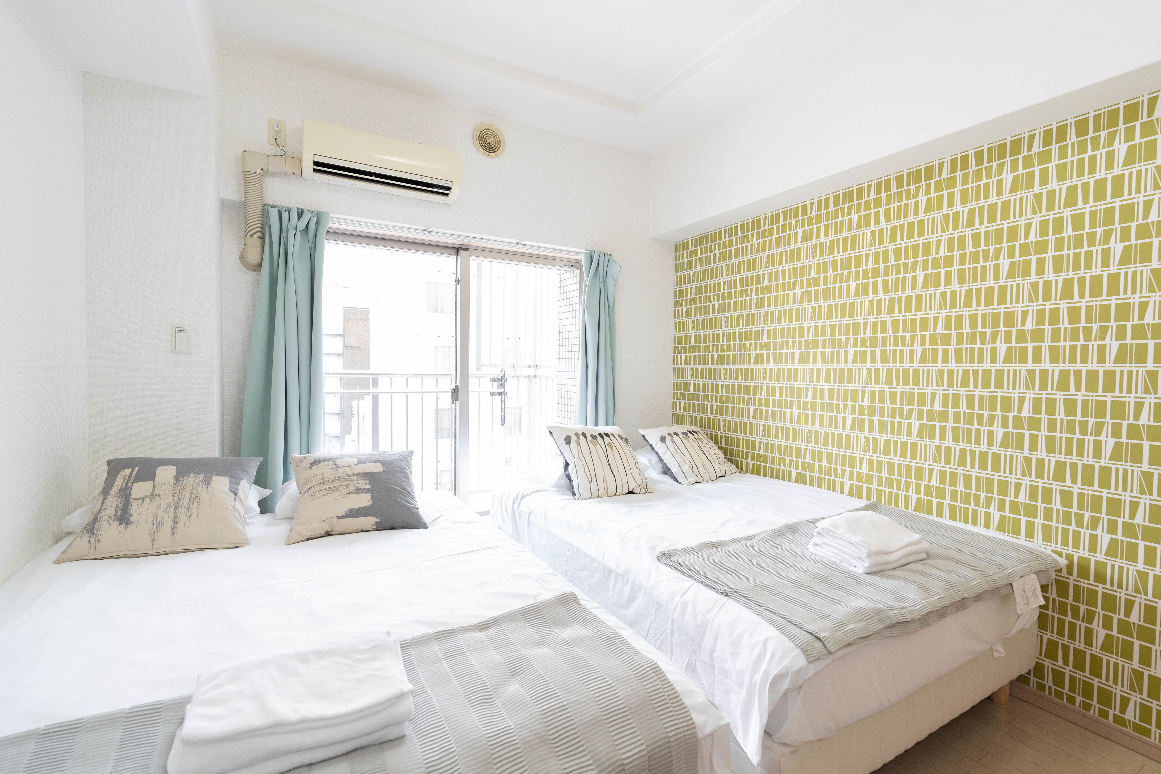 A hotel room with two beds, a window with curtains, an air conditioning unit, and a green mosaic wall.