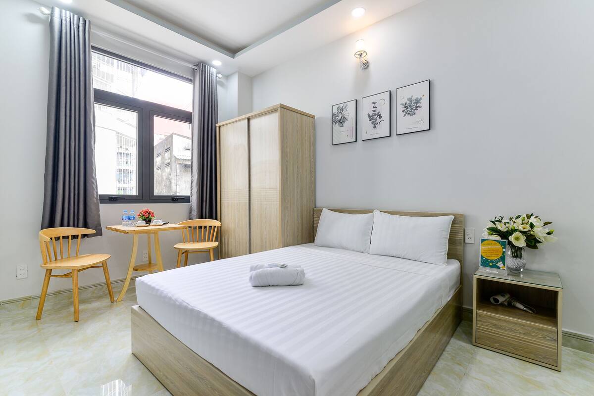 Une chambre d’hôtel avec un lit, deux chaises en bois, une petite table, une armoire et une table de chevet ornée d’un vase de fleurs.