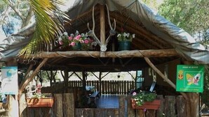A rustic wooden structure with a thatched roof, surrounded by potted plants and a wooden fence. - La Petite Ferme (La Foa)