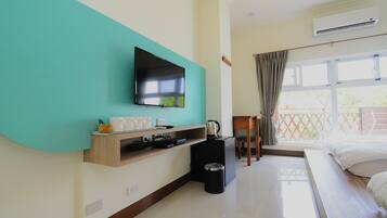 A hotel room with a flat-screen TV mounted on a teal wall, a small kitchenette with a coffee maker, a trash bin, a wooden chair, and a bed with white linens.