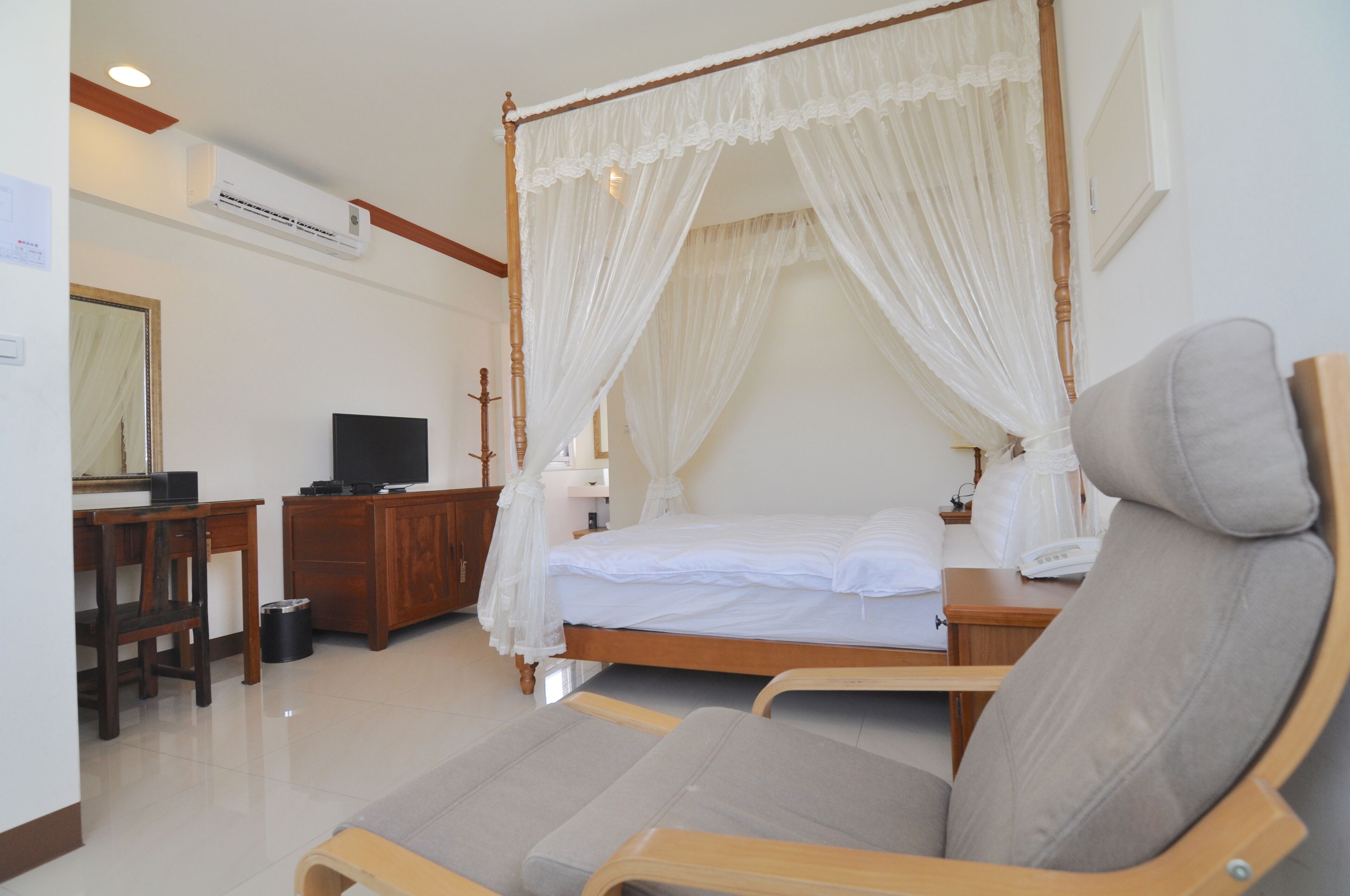 A four-poster canopy bed with a wooden frame and white bedding, a television on a wooden stand, a desk with a chair, and a trash can.