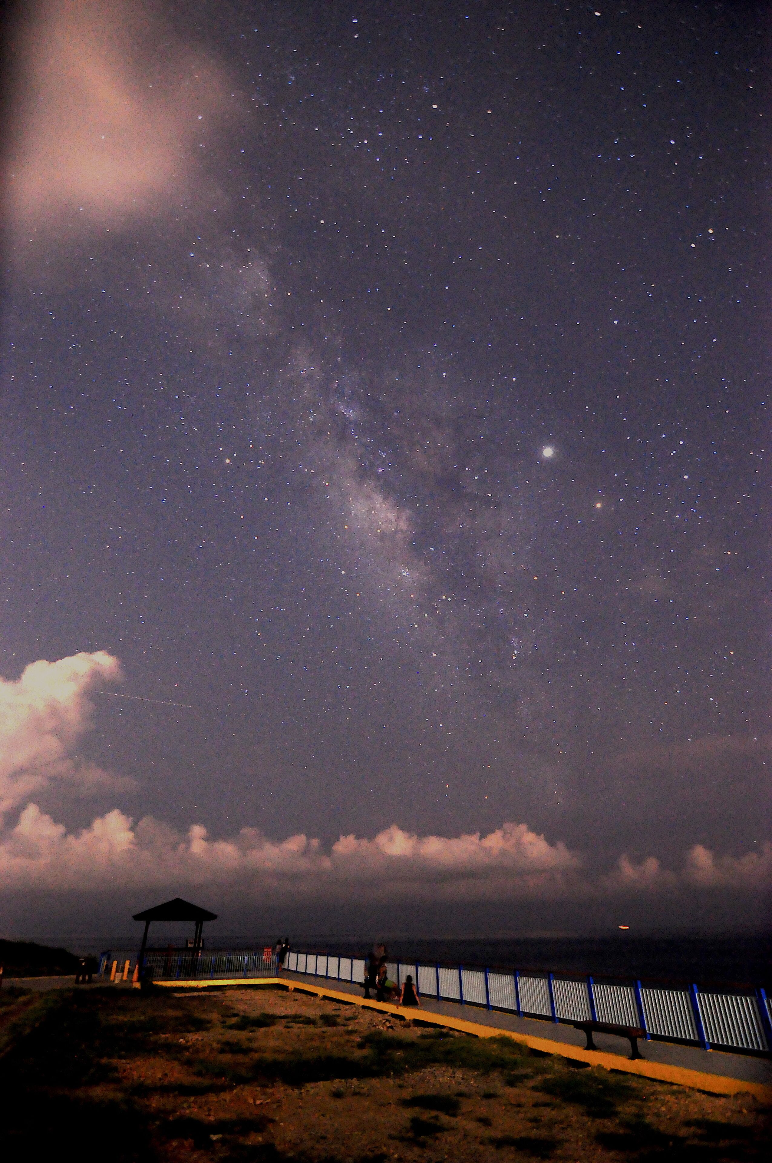 A night sky with stars and the Milky Way, a walkway with a railing, and a small shelter.