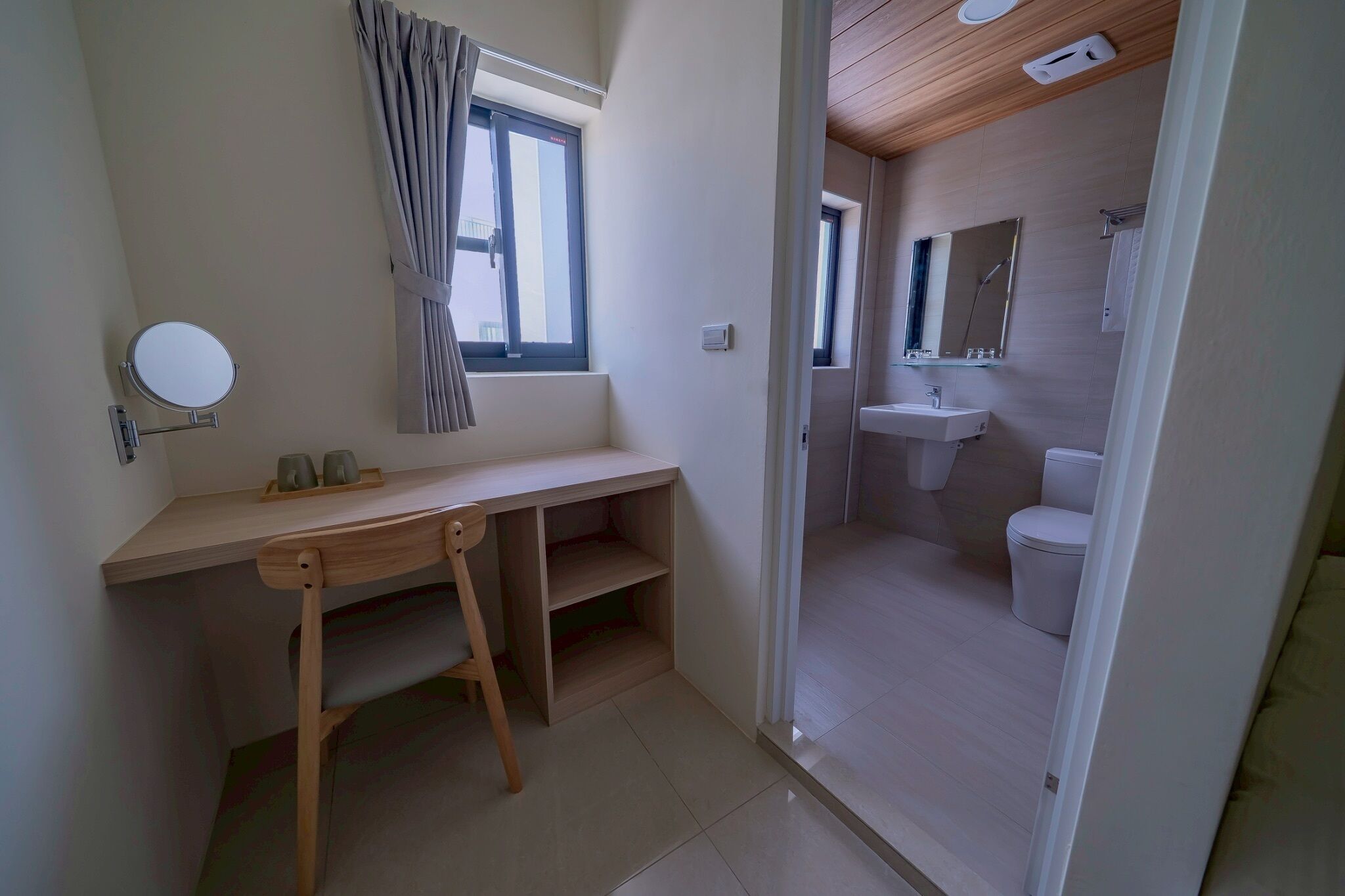 A small, clean bathroom with a toilet, sink, and mirror. There is a window with curtains, a small desk with a mirror, and a shelf.