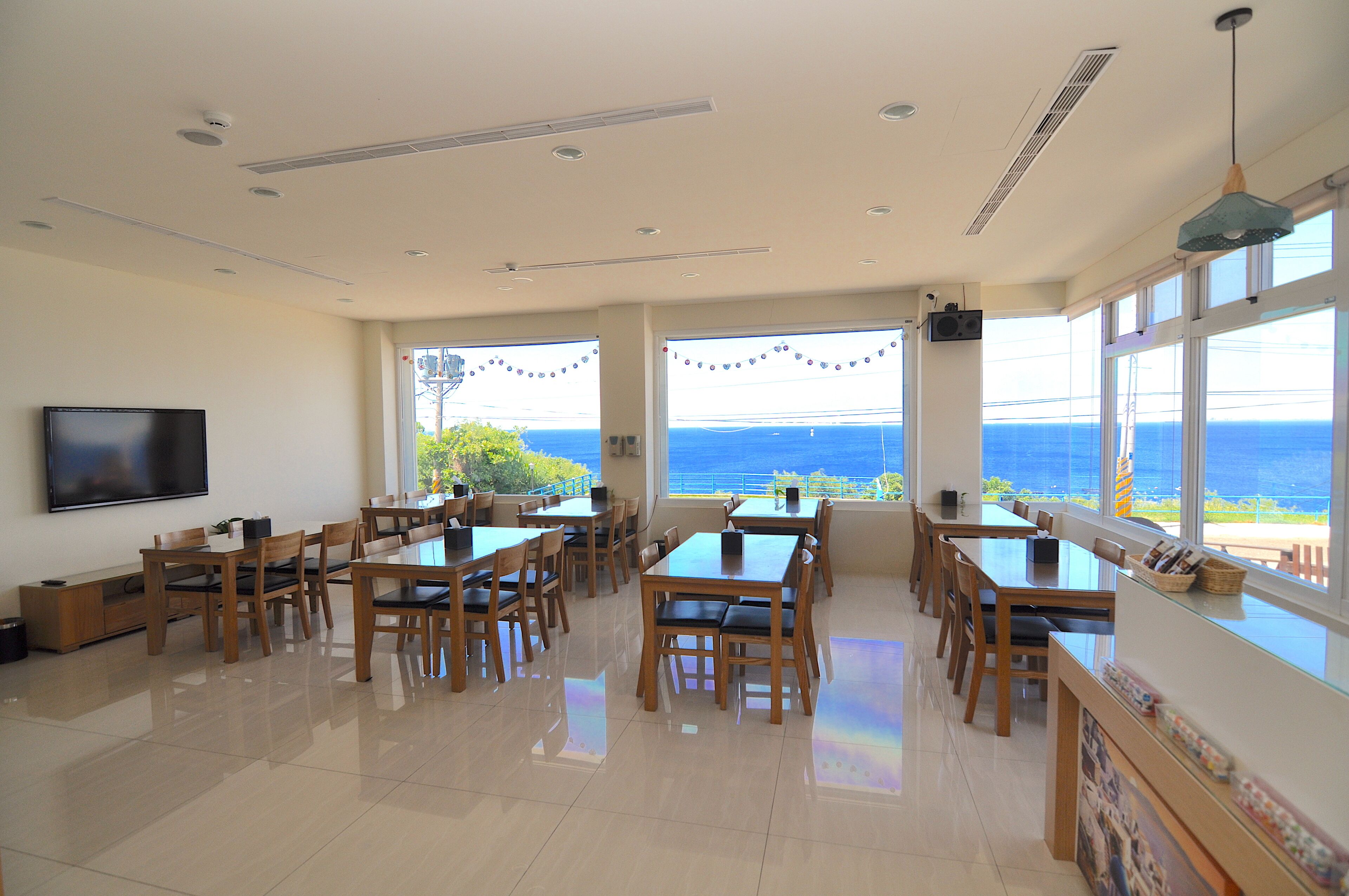 A spacious dining area with multiple wooden tables and chairs, large windows offering a view of the sea, and a flat-screen TV mounted on the wall.