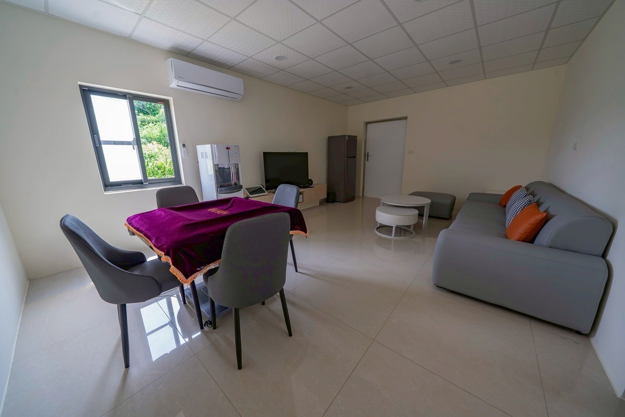 Una sala de estar moderna con comedor, un sofá gris, un televisor y un refrigerador.