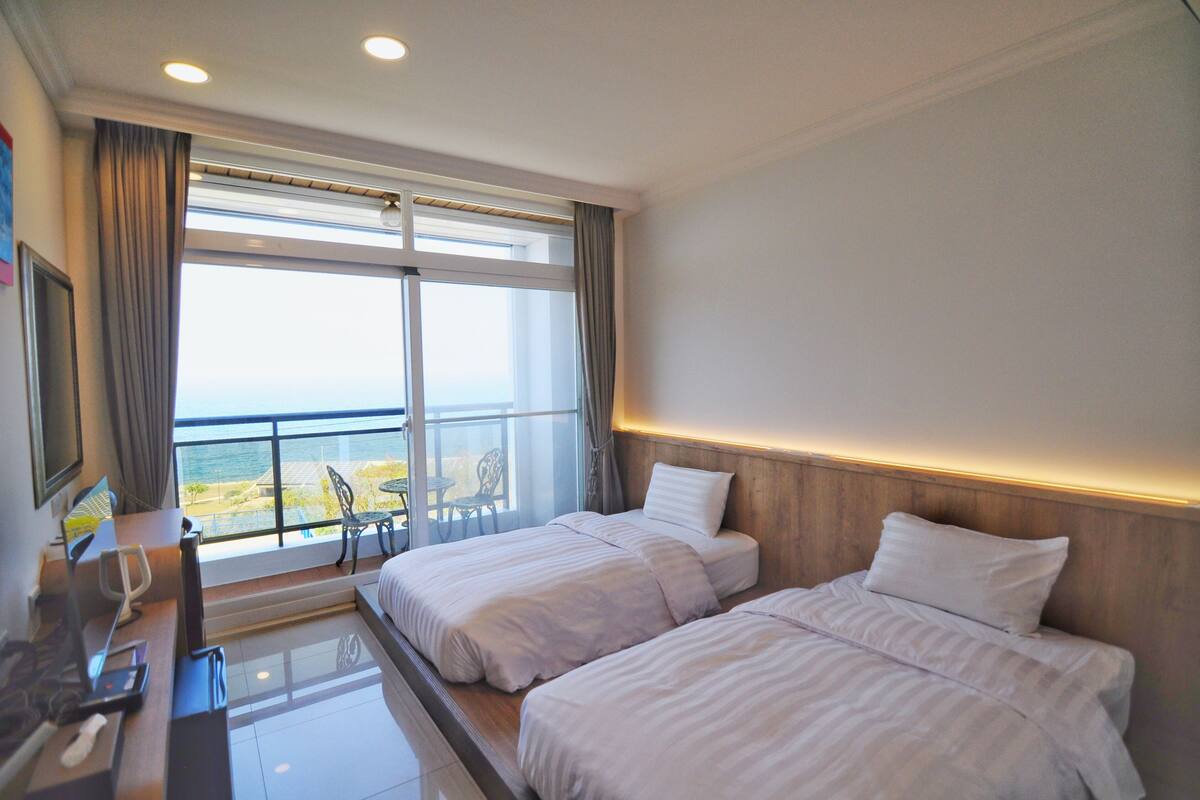 A hotel room with two beds, a desk with a telephone, and a view of the sea through large windows.