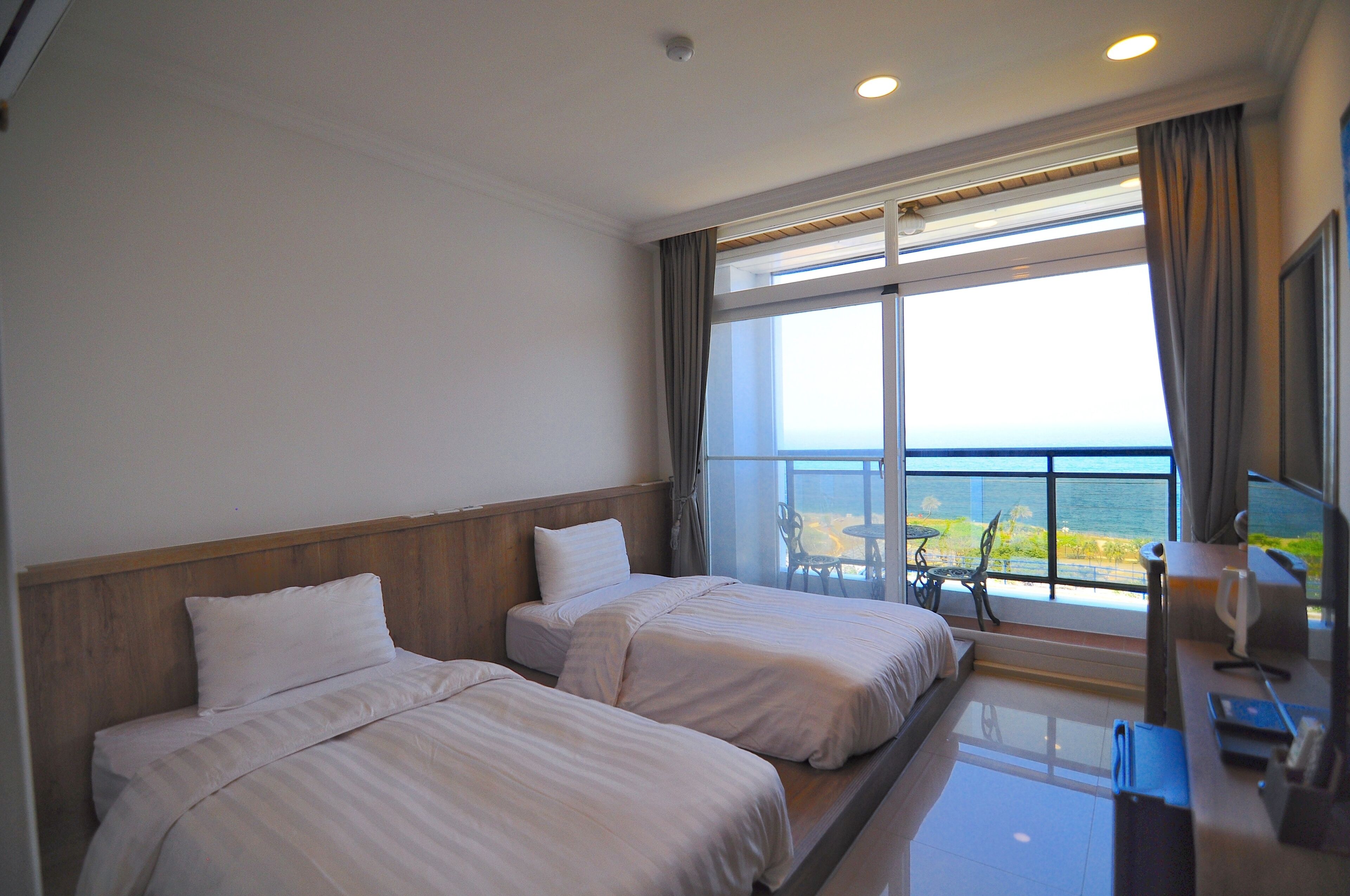 A hotel room with two beds, a desk with a chair, a window with a view of the sea, and a balcony with outdoor seating.