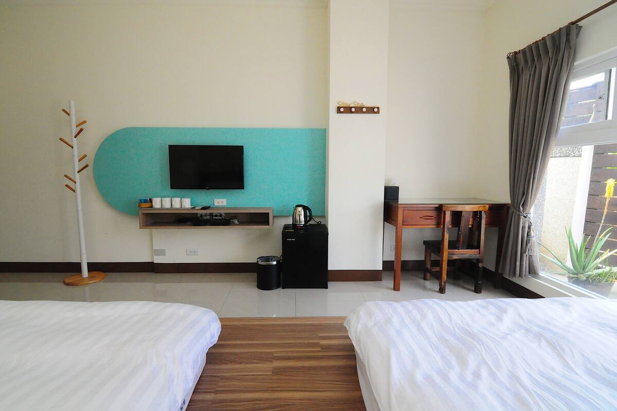 A hotel room with two beds, a desk, a television, a small table, a trash bin, and a window with curtains.