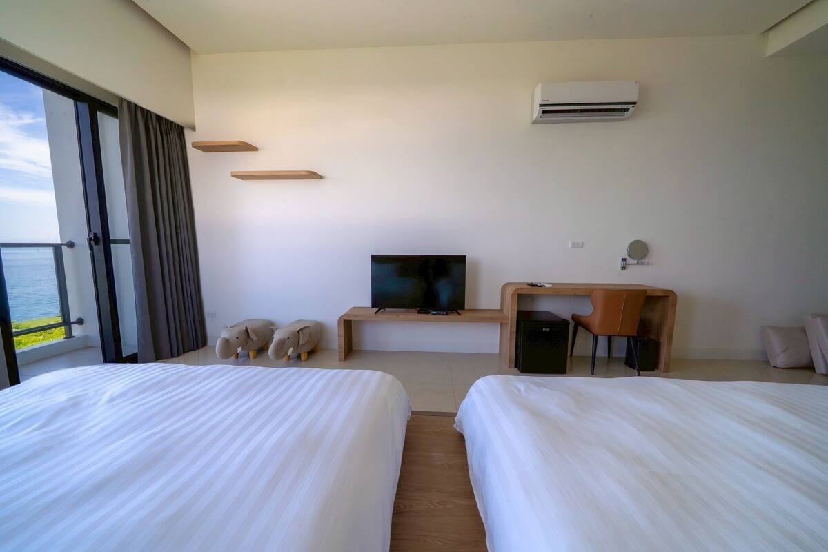 A hotel room with two beds, a TV, a desk with a chair, and a view of the sea.