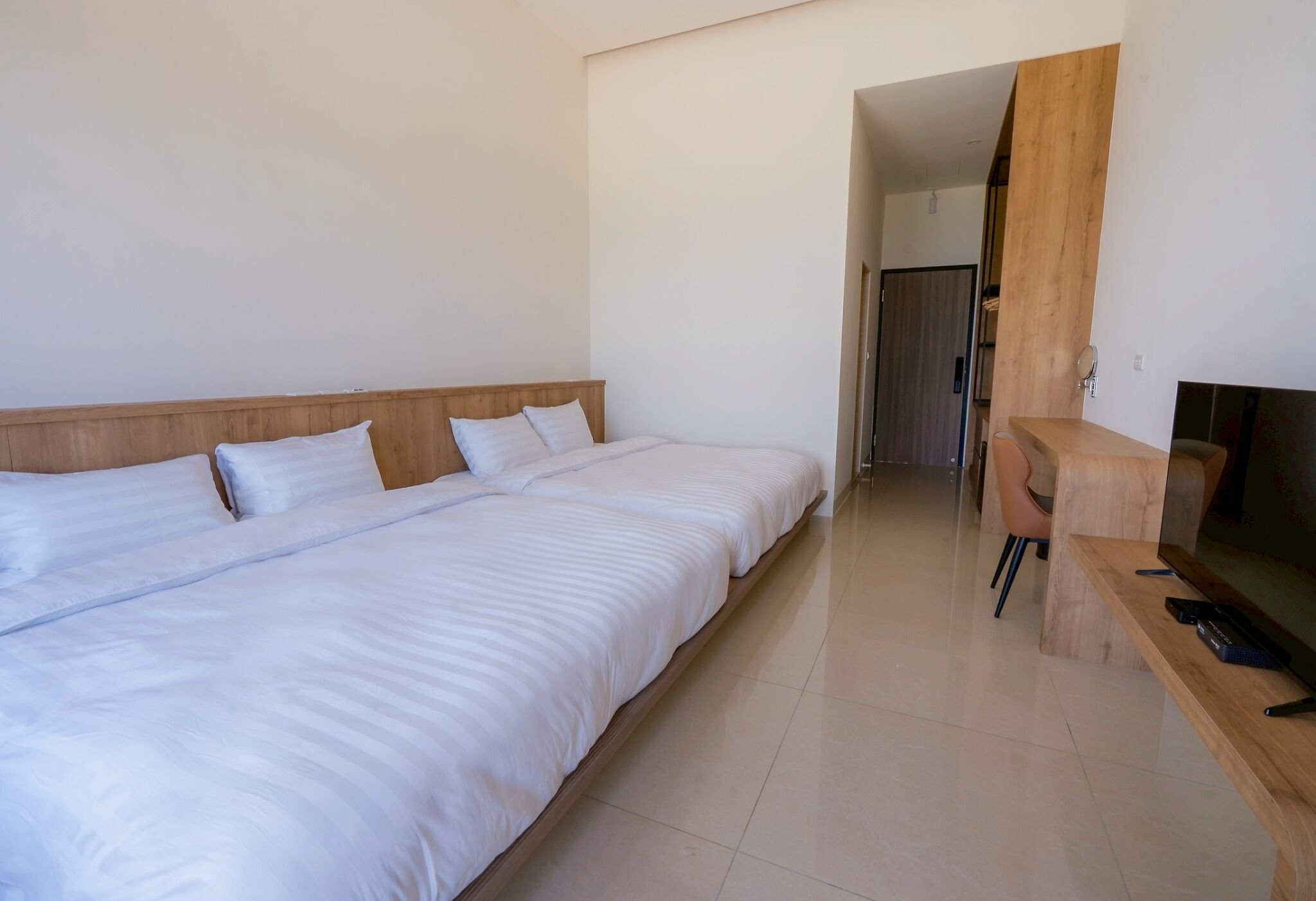 Habitación de hotel con dos camas individuales, cabecero de madera, escritorio de madera con televisión y una puerta que da a otra habitación.