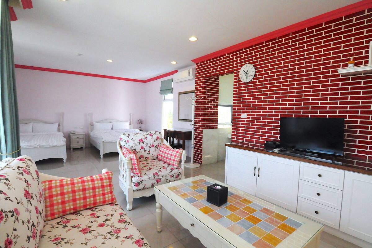 A hotel room with two beds, a floral sofa, a television, and a red brick accent wall.