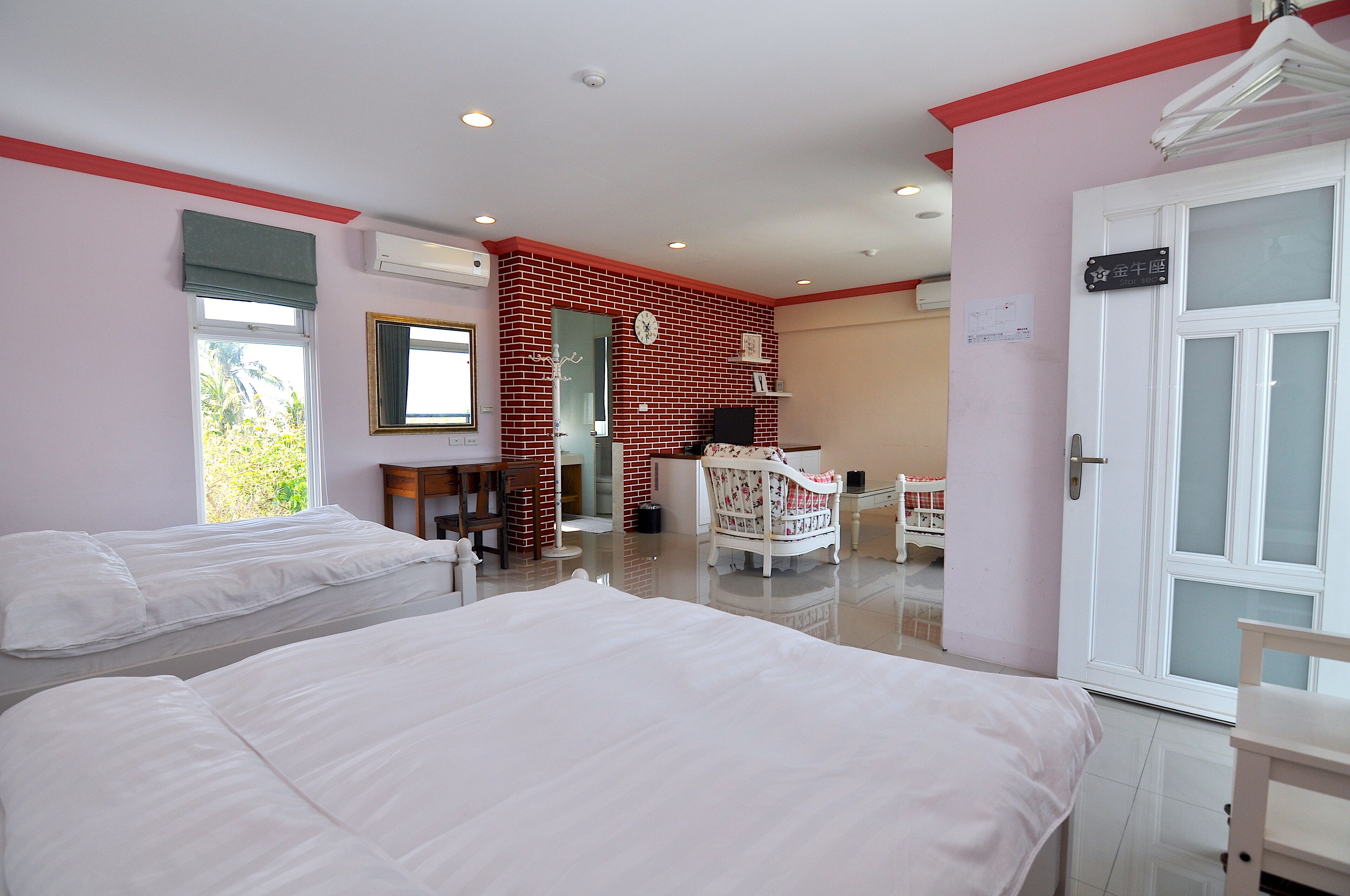 A hotel room with two beds, a sitting area with a chair and table, a mirror, and a door with a sign.