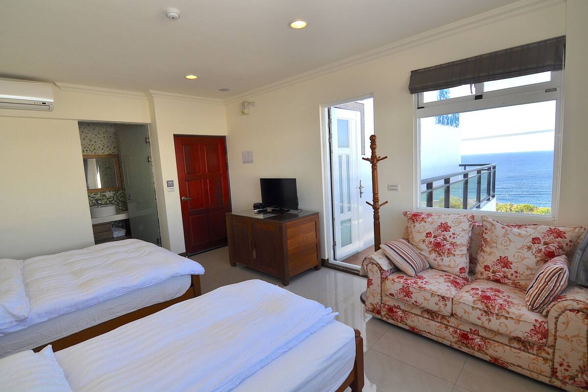 A hotel room with two beds, a sofa, a television, and a view of the sea.