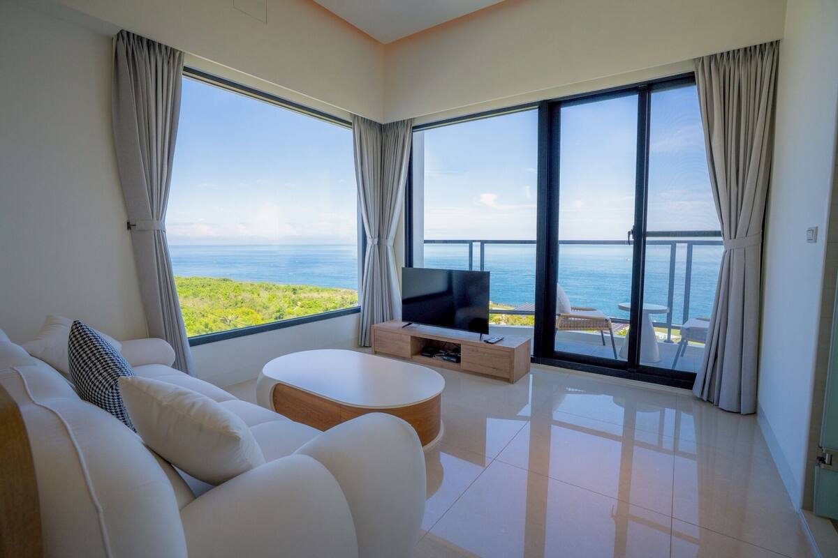 A modern living room with a large TV, white furniture, and a balcony with a view of the sea.