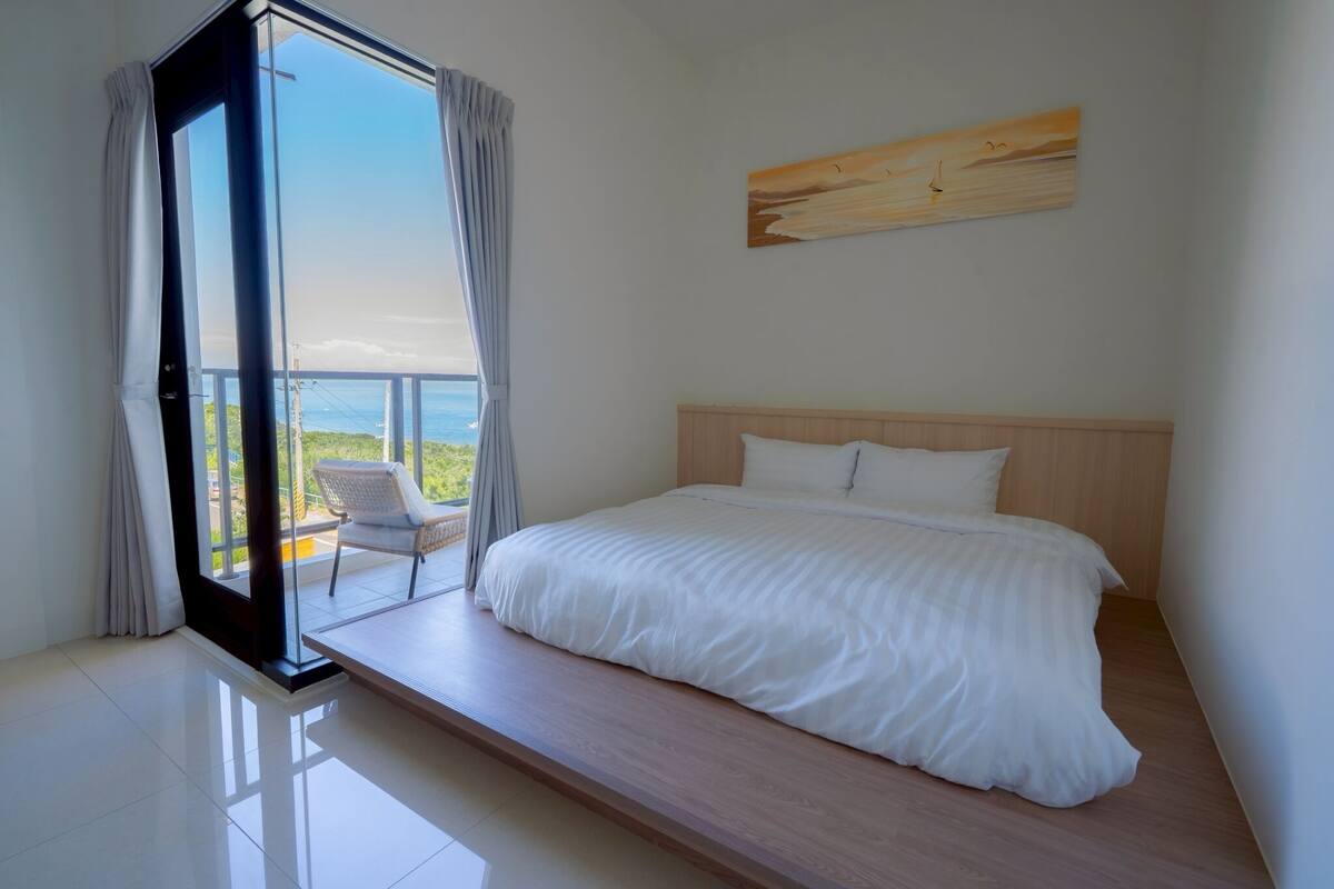 A bedroom with a large bed, a view of the sea through a sliding glass door, and a wooden headboard.