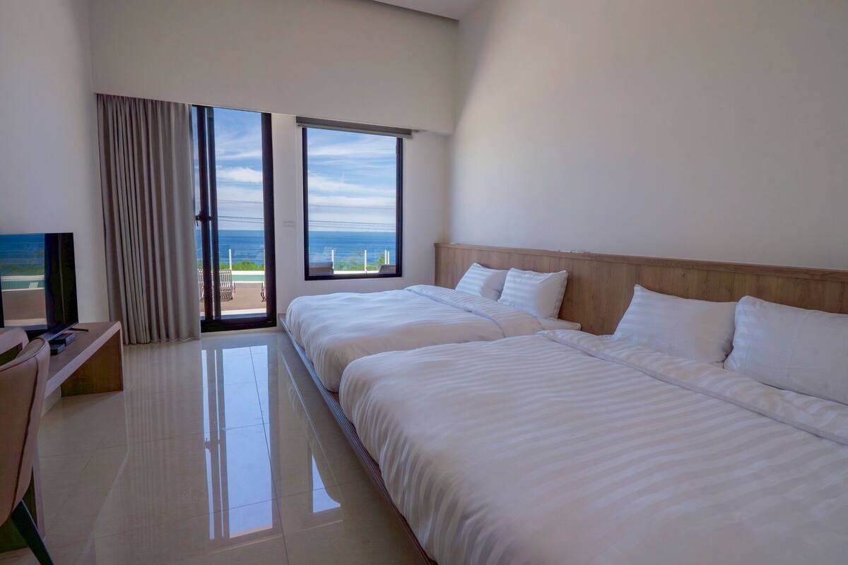 A hotel room with two beds, a TV, and a balcony with a view of the ocean.