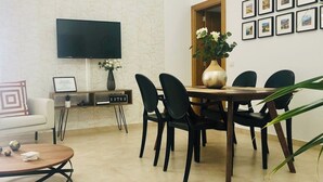Un salon moderne avec un téléviseur à écran plat, une table basse, un canapé, une table à manger avec des chaises, un vase de fleurs et une étagère murale ornée d’un panneau d’accueil encadré et d’un routeur.