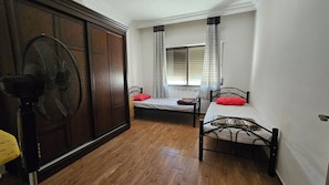 A room with two single beds, a wooden wardrobe, a standing fan, and a window with patterned curtains.