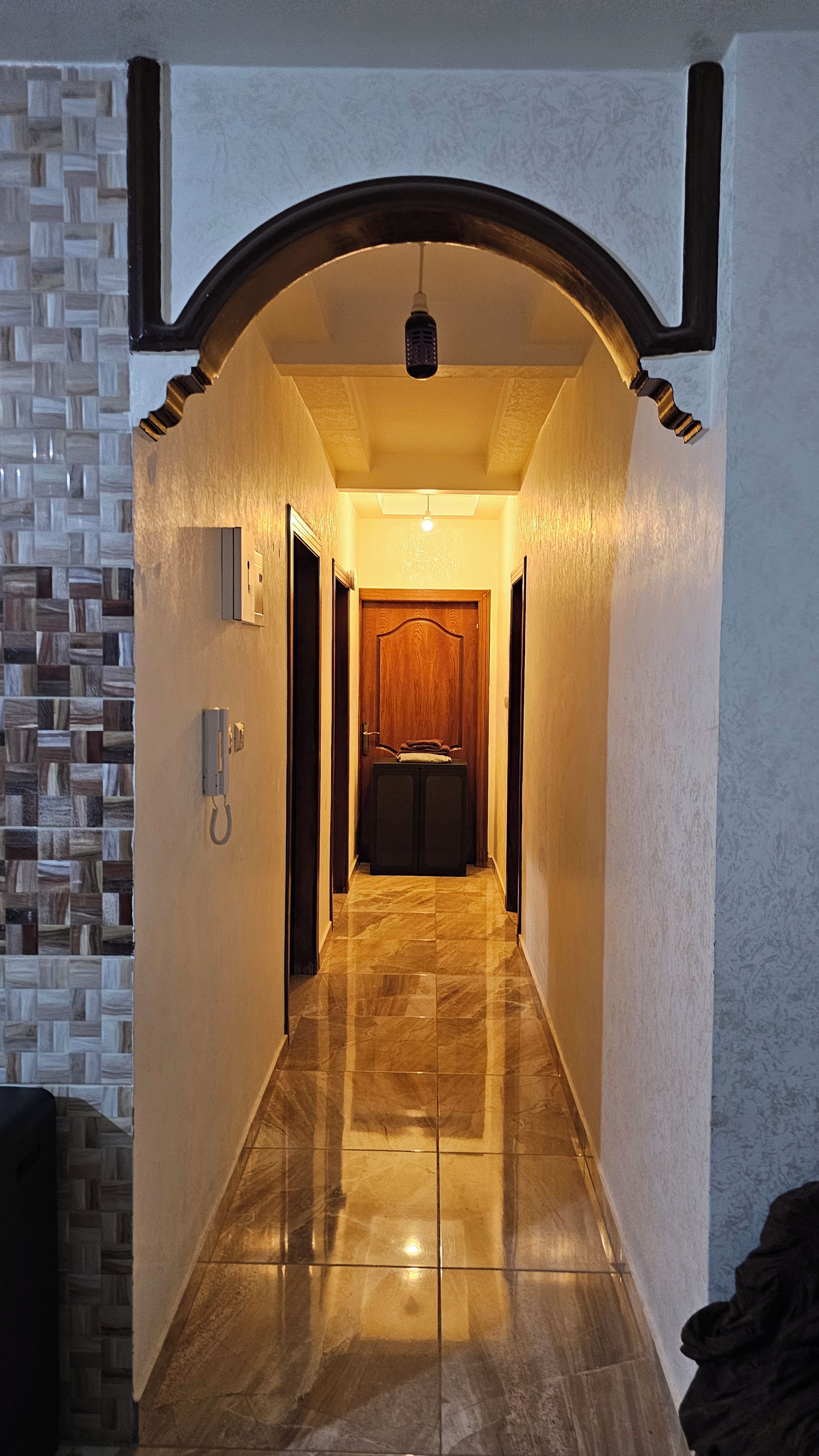 A hallway with a wooden door at the end, a telephone on the wall, and a patterned wall on the left.