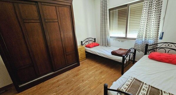 A bedroom with a wooden wardrobe, a single bed with a red pillow, and a window with patterned curtains. - Private Family Apartment - Very Quiet Location (Amman)