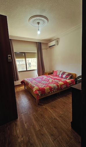 Quarto com cama de madeira, ventilador de teto, ar condicionado, janela com persianas e vista para prédios.