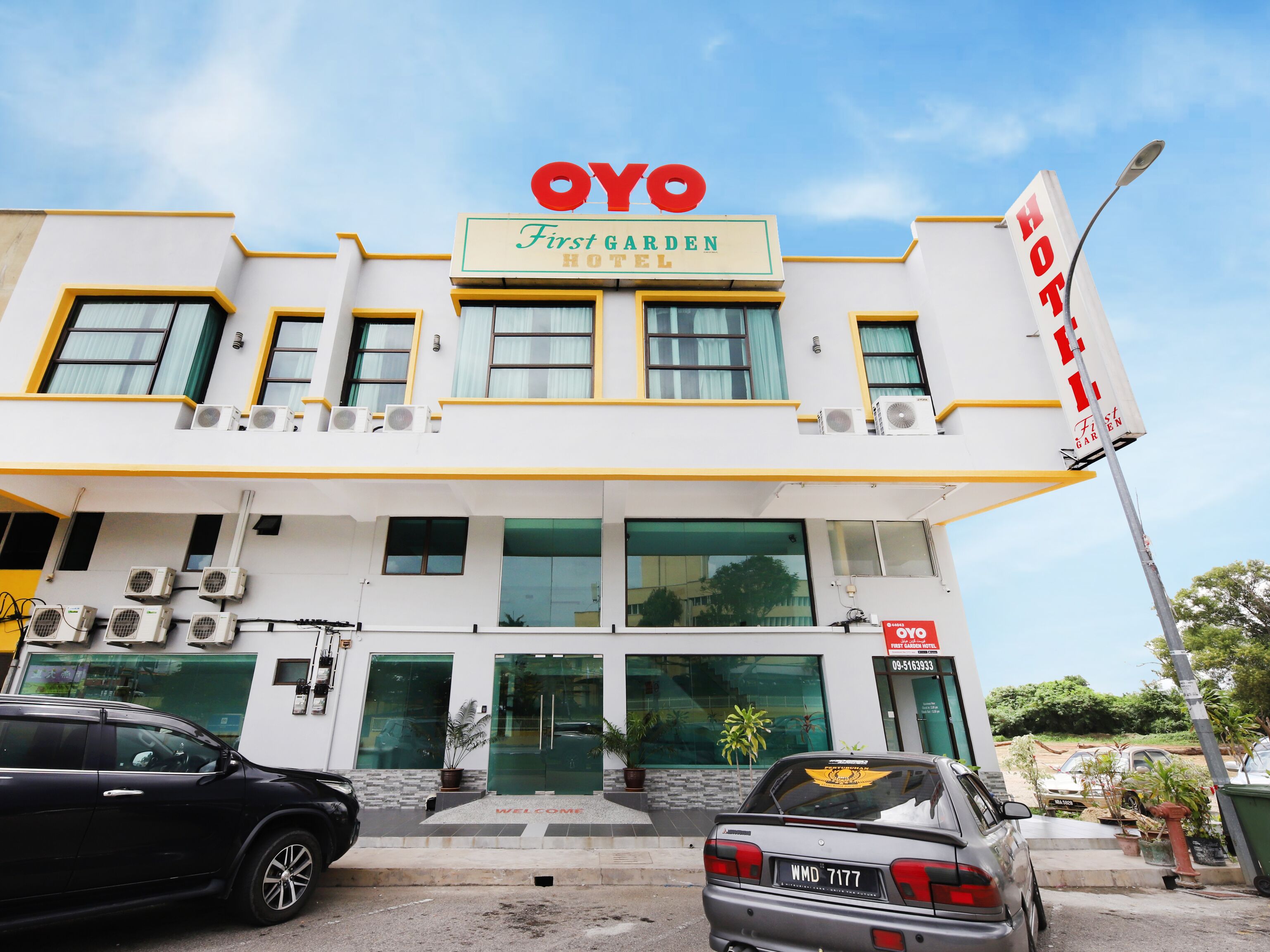 Ein Hotel namens 'OYO First Garden Hotel' mit modernem Design, das mehrere Klimaanlagen, einen einladenden Eingang und eine gut sichtbare Kontaktnummer zeigt.