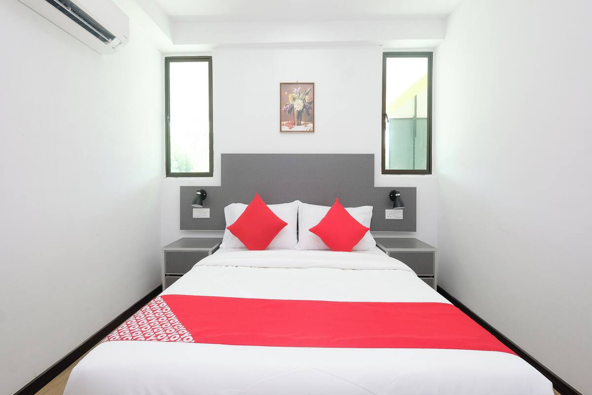 A hotel room with a bed, two bedside tables, a nightstand, a wall-mounted lamp, and a window.