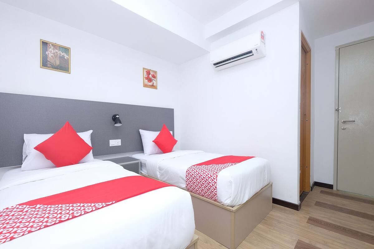 A hotel room with two beds, each with white bedding and red accents, a grey headboard, a bedside lamp, and an air conditioning unit on the wall.