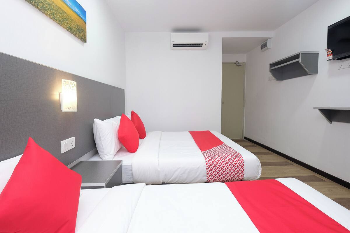 A hotel room with a bed, red and white bedding, a grey headboard, a wall-mounted air conditioner, a painting, and a small shelf.
