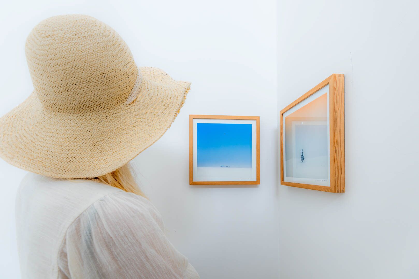 A person wearing a straw hat and a light-colored shirt, standing in front of two framed artworks on a white wall.