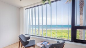 Habitación con un ventanal grande con vista a la playa, dos sillas grises y una mesita con un libro.