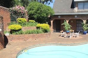 A brick building with a swimming pool, surrounded by well-maintained hedges and potted plants.