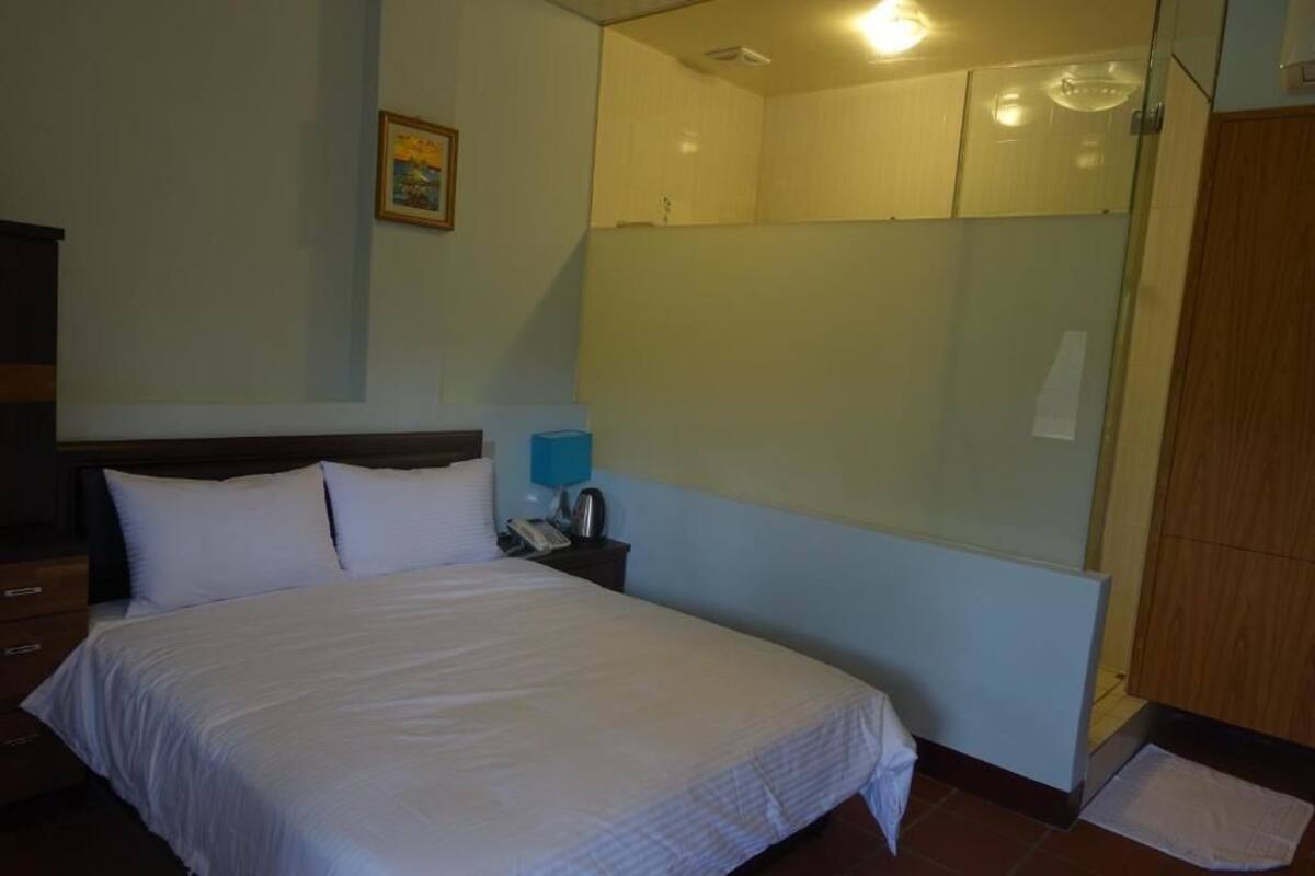 Habitación de hotel con cama, mesita de noche, lámpara azul, cuadro en la pared y zona de ducha.