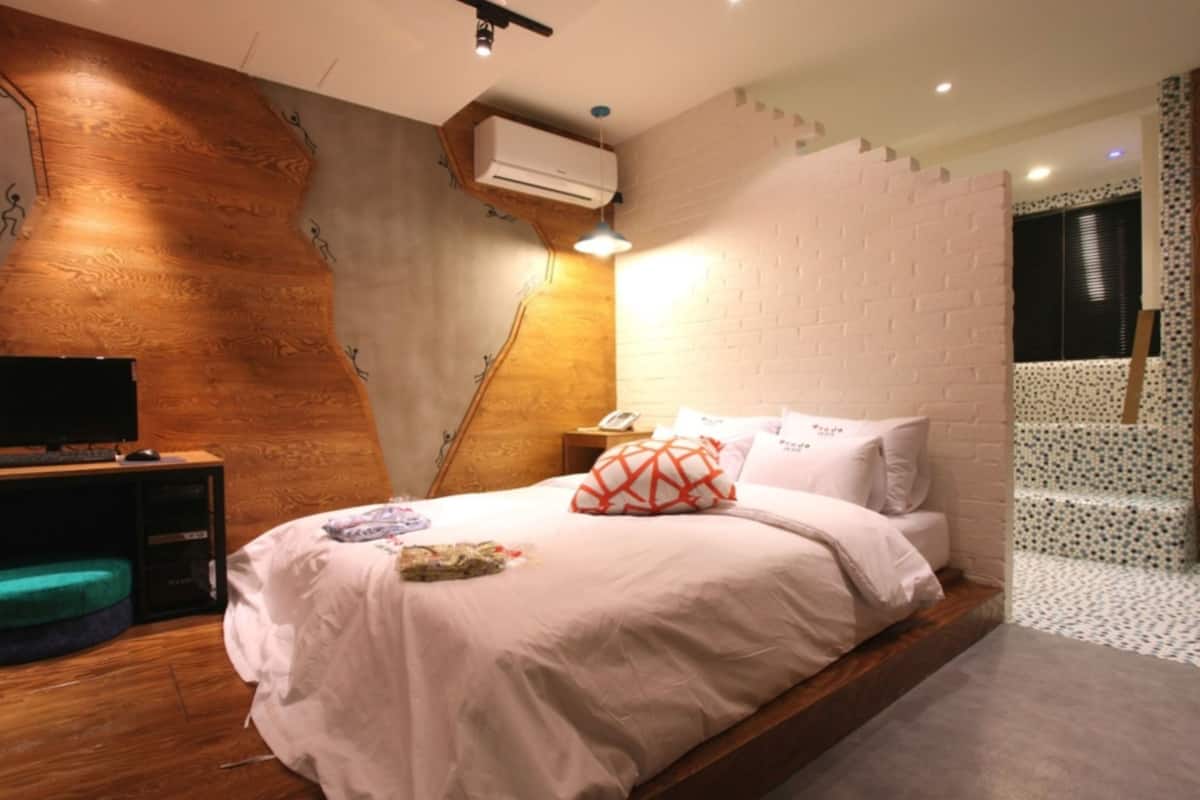 A bedroom with a wooden accent wall, a bed with white bedding, a desk with a computer, and an air conditioning unit.