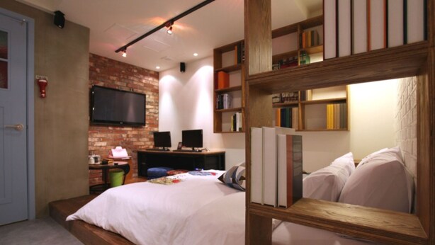 A bedroom with a brick wall, a flat-screen TV, a bookshelf, and a bed with white bedding. - Hotel Yaja Suwon City Hall (Suwon)