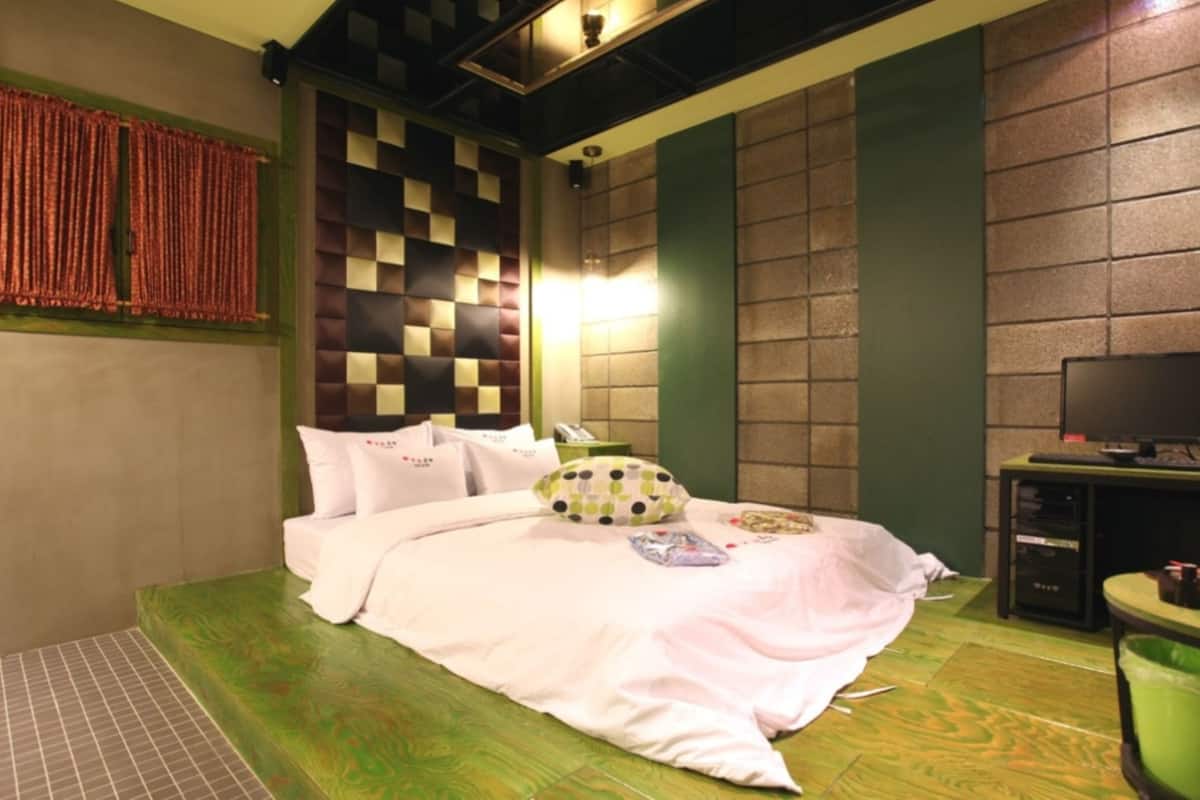 A neatly made bed with pillows and a blanket, a television on a stand, and a green trash bin.