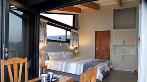 A bedroom with a bed, a wooden table, a chair, a window with curtains, a bedside lamp, and a wardrobe. - Forever View (Knysna)