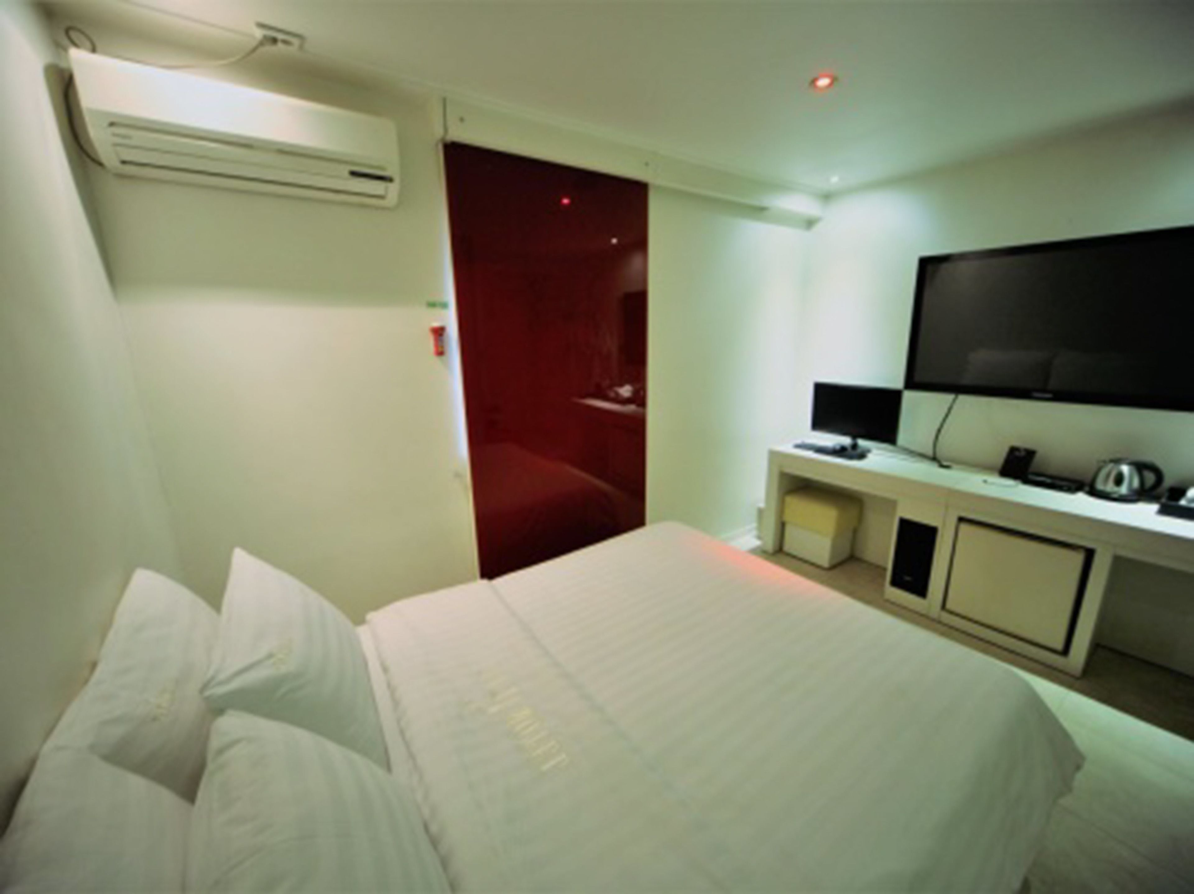 A hotel room with a bed, a TV, a desk with a computer, and an air conditioning unit.
