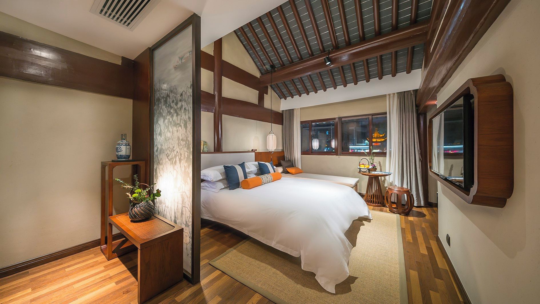 A hotel room with a large bed, a bedside table with a vase, a window with a view of a building, and a wooden ceiling.