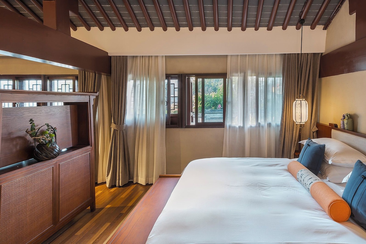 A hotel room with a large bed, a wooden headboard, a nightstand, a potted plant, and a window with curtains.