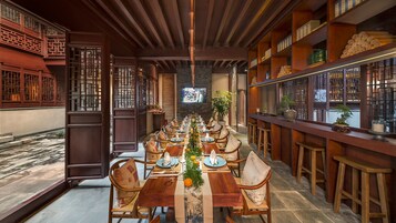 Un restaurante chino tradicional con una larga mesa de comedor de madera dispuesta para una comida, rodeada de sillas de madera y una zona de bar con estanterías.