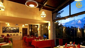 A restaurant with multiple tables set for dining, large windows offering a view of the outdoors, and a ceiling with exposed beams and hanging lights. - Queens Meadows by Sunshine & Smiles (Ranikhet)
