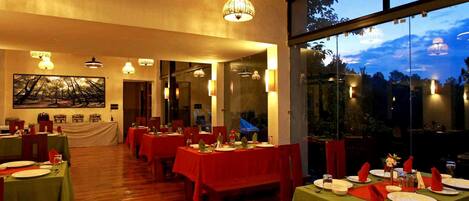 A restaurant with multiple tables set for dining, large windows offering a view of the outdoors, and a ceiling with exposed beams and hanging lights.