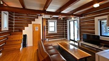 A log cabin interior with a wooden staircase, a sofa, a dining table, and a television.