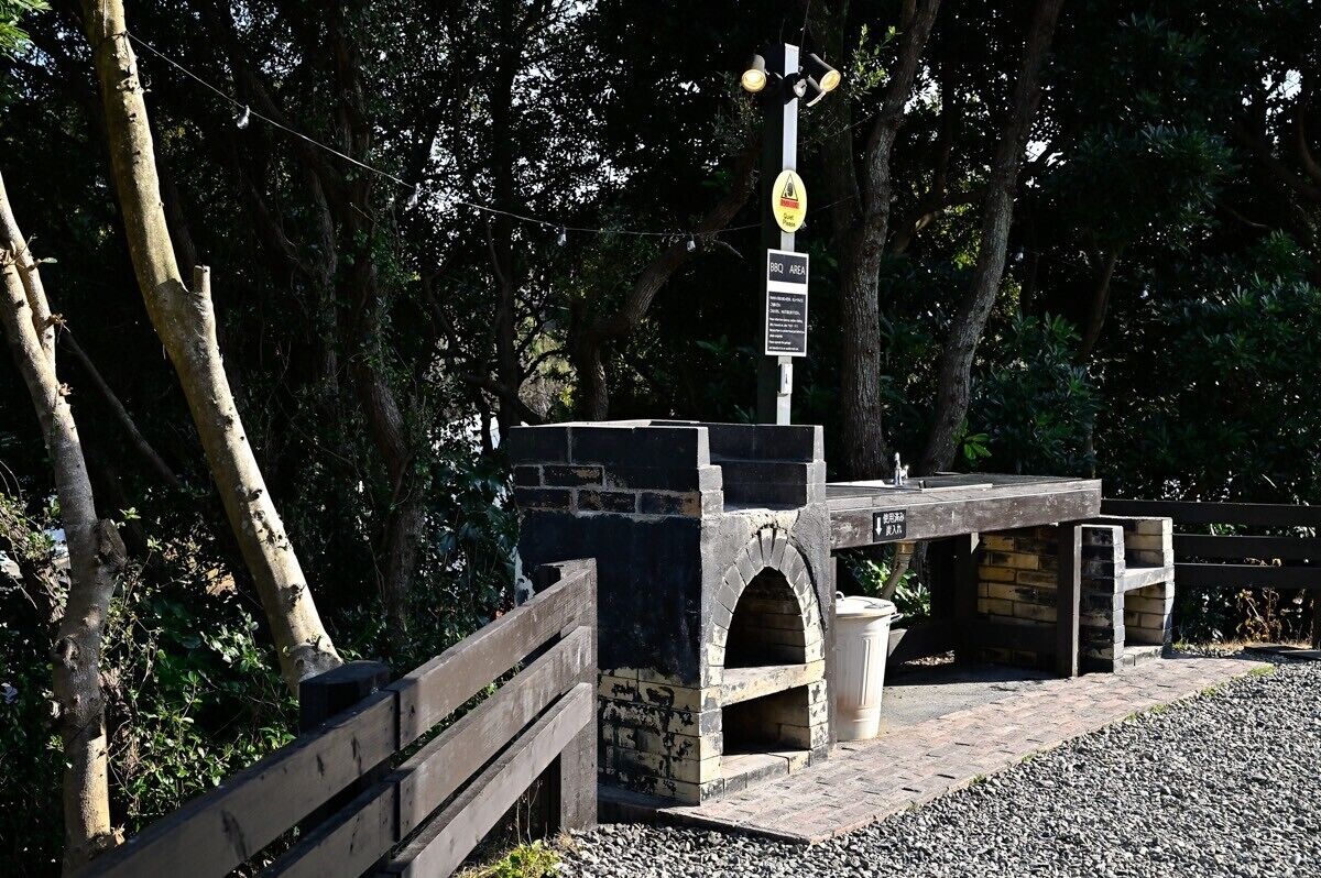 Zona de barbacoa de piedra con horno de ladrillo, rodeada de árboles y una valla de madera.