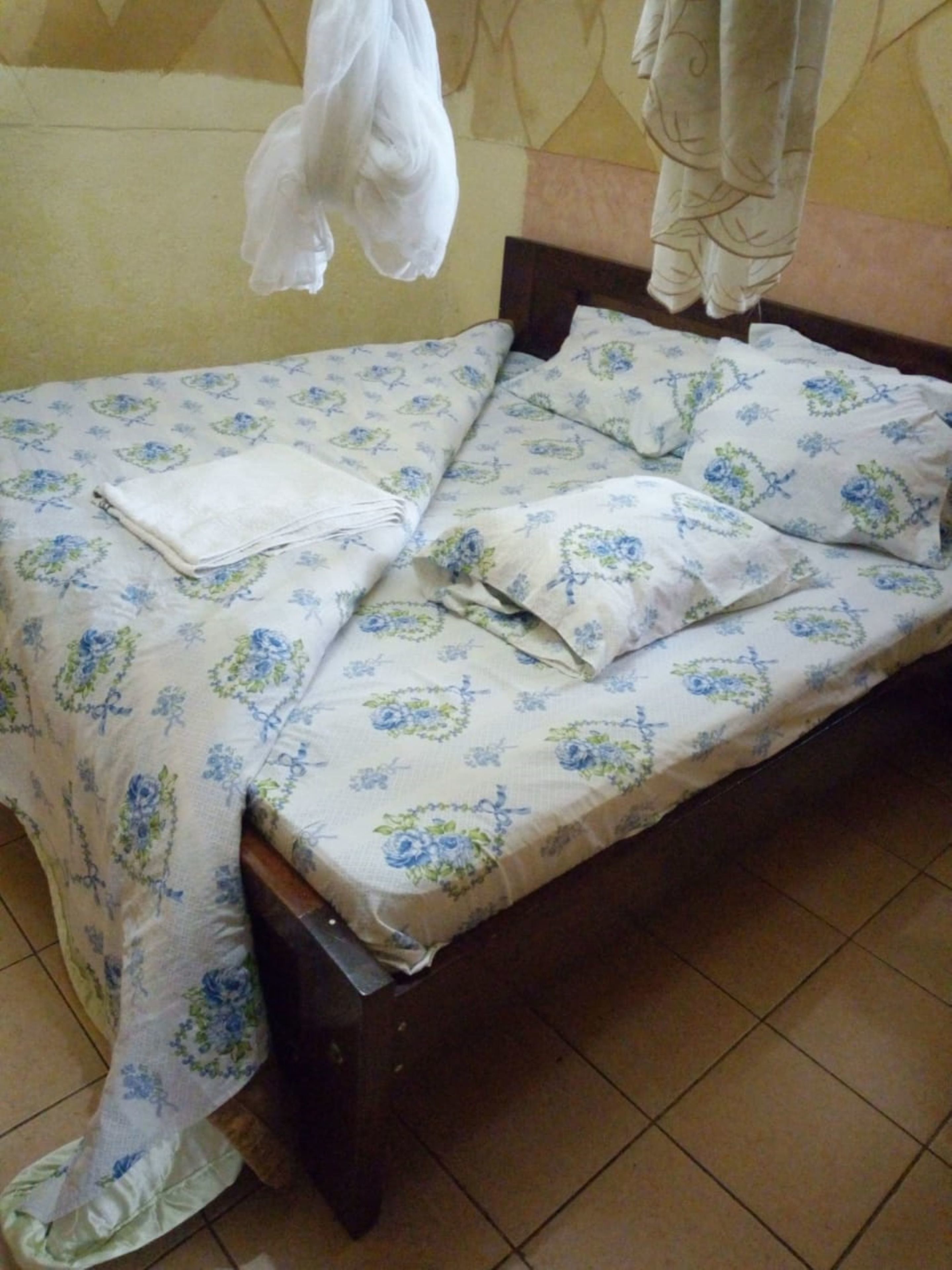 A small, single-bed room with a floral-patterned bedspread, a wooden headboard, and a tiled floor.