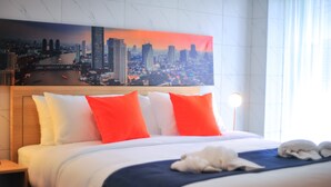 Habitación de hotel con cama, almohadas, una manta, un cuadro de un paisaje urbano y una mesita de noche.