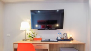 Habitación de hotel con un escritorio de madera, una silla roja, un televisor montado en la pared, una lámpara, una planta pequeña, una caja de pañuelos, una bandeja con botellas y una cafetera, y un refrigerador pequeño.