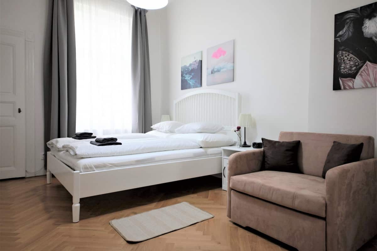 A bedroom with a white bed, a beige sofa, and a wooden floor.